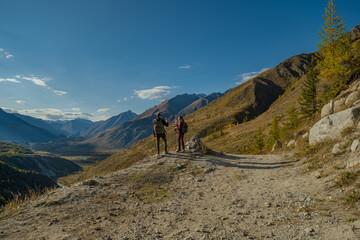 Fototapeta premium Couple of hikers with backpacks standing on edge of mountain road looking at scenic alpine valley during autumn trekking trip for travel adventure, partnership and freedom concept.