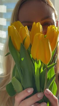 Smiling blonde woman smelling a fresh bouquet of yellow tulips, happily enjoying the floral aroma and spring sunshine coming from the window, expressing pure delight and serenity
