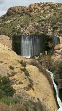 Water cascading over the elche reservoir dam, a historic gravity dam surrounded by rocky mountains and green vegetation under a cloudy sky in alicante, spain