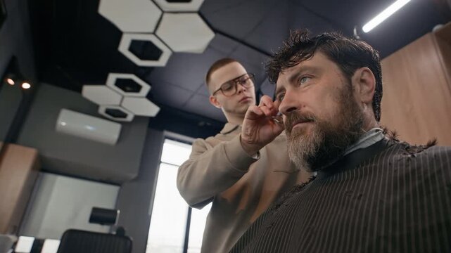 Low angle shot of young Caucasian barber standing behind middle aged Caucasian client, cutting his hair with comb and scissors