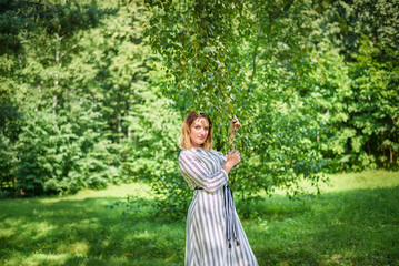 Naklejka premium A woman in a white dress stands in a sunlit forest clearing on a summer day. In front of her is lush green grass, and behind her are tall, slender trees.