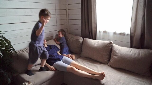 Two boys walking and stepping over relaxed mother on couch with dog nearby, midday light and gentle disorder, scene captures childhood independence, boundary testing and family rhythm as symbolic rite