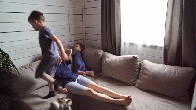 Playful boys happily jump on the couch. around a tired mom, while mom is lying on the couch. Children do not pay attention to their mother's fatigue and play pranks