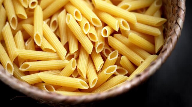 Dry wheat penne pasta in rustic wicker basket close up on dark background. Uncooked Italian ingredient texture