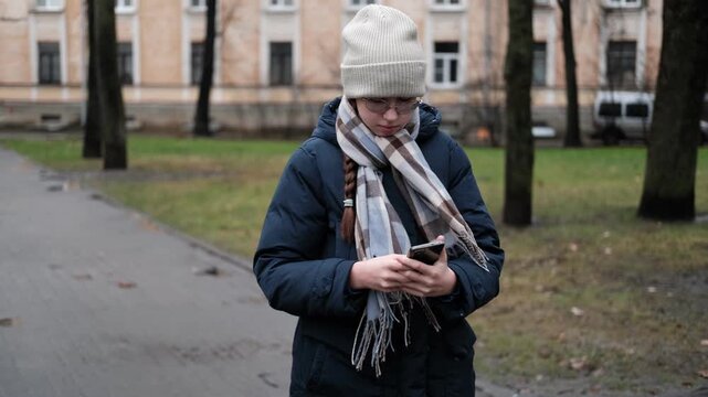 Teenager Girl Walking Alone on City Street with Smartphone. Portrait of 14 years old girl wearing eyeglasses, wool hat, winter jacket using mobile phone. Urban Style. Rainy mood weather. Cold season
