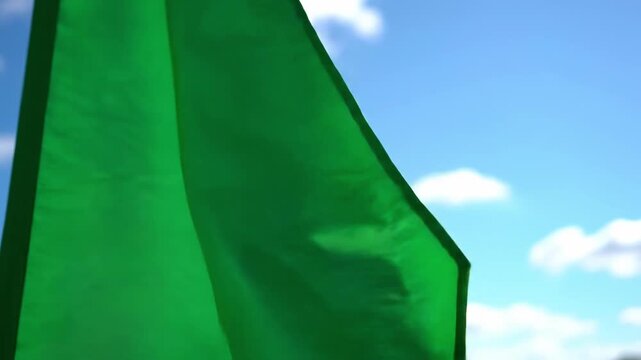 A vibrant green flag flutters energetically against a bright blue sky with fluffy clouds, evoking feelings of hope, progress, and positive change. Ide