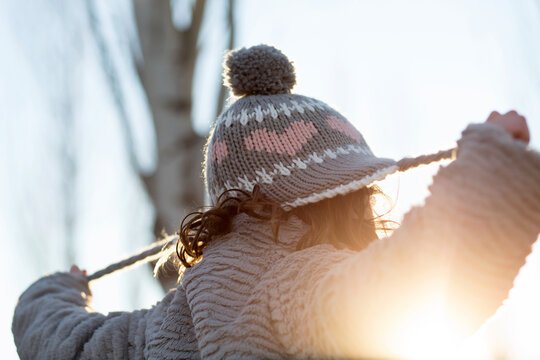 Rear view of a girl in warm clothing standing in a garden pulling the ties on her woolly hat