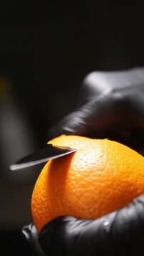 cocinero profesional con guantes de l&aacute;tex negro pela cuidadosamente una naranja fresca, utilizando movimientos controlados del cuchillo para retirar la piel de forma limpia y preparar la fruta.