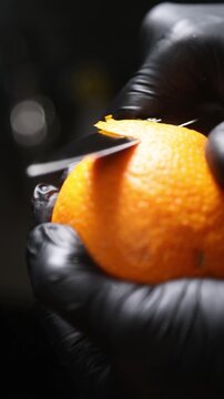 cocinero profesional con guantes de l&aacute;tex negro pela cuidadosamente una naranja fresca, utilizando movimientos controlados del cuchillo para retirar la piel de forma limpia y preparar la fruta.