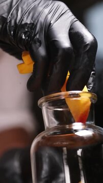 Un cocinero profesional con guantes de l&aacute;tex negro introduce trozos de melocot&oacute;n y tiras de piel de naranja en un bote de cristal transparente, preparando los ingredientes para elaborar un alm&iacute;bar