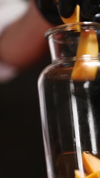 Un cocinero profesional con guantes de l&aacute;tex negro introduce trozos de melocot&oacute;n y tiras de piel de naranja en un bote de cristal transparente, preparando los ingredientes para elaborar un alm&iacute;bar