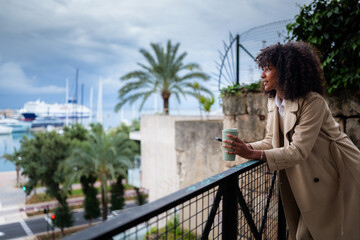 Young black woman leans on balcony railing with reusable coffee cup and smartphone, gazing over a...