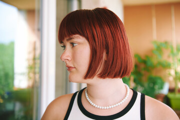 Side profile portrait of teenage girl with red hair and pearl necklace looking away outdoors near window and green plants, natural youth lifestyle photo © annanahabed