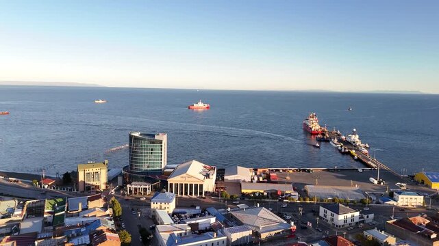 Aerial footage moving forward from the colorful rooftops of Punta Arenas toward the Strait of Magellan, featuring the Dreams Casino tower, Prat Pier, and ships anchored in the blue waters during sunse