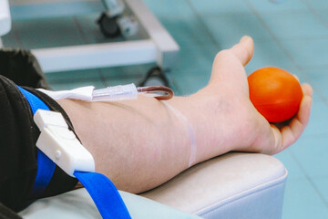 Human arm with intravenous line and blood donation equipment holding an orange fruit, seated on a medical chair in a healthcare facility