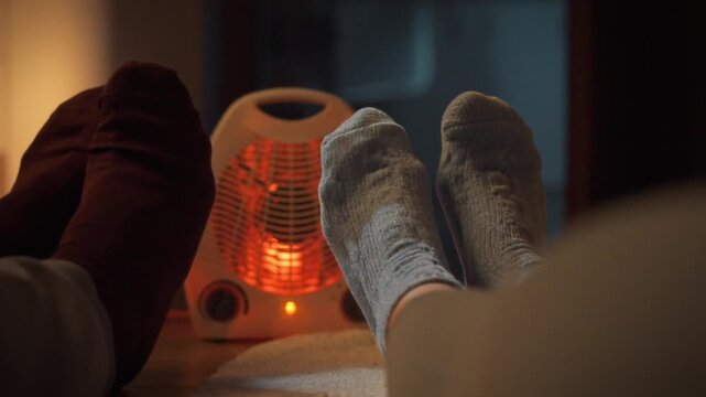 Cozy winter evening scene unfolds as young couple relaxes on the floor, clad in warm socks, basking in the warmth of heater that casts bright red glow on their feet, fostering intimacy and relaxation