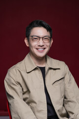 Obraz premium Smiling Asian man wearing glasses sitting against a red studio background. Confident male portrait with friendly expression, modern lifestyle concept, professional look, positive emotion, and clean 