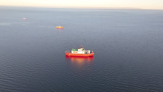 Aerial moves toward the orange-hulled Almirante Viel in the Strait of Magellan, tilting down to reveal the green deck and circular helideck during a golden sunset in Punta Arenas