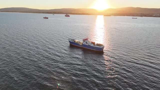 Aerial circling the Chilean wellboat Calipso near Punta Arenas at sunset, highlighting its blue and white hull against the golden reflection on the Strait of Magellan