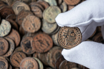 Hand in white glove holding ancient Roman coin of Maximianus. Numismatist examining bronze emperor portrait coin from archaeological hoard. History and archaeology concept © scaliger