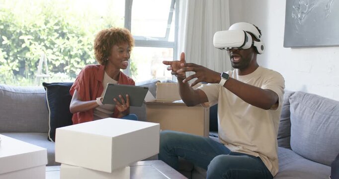 African American couple sitting on couch unpacking boxes man testing VR headset woman using tablet