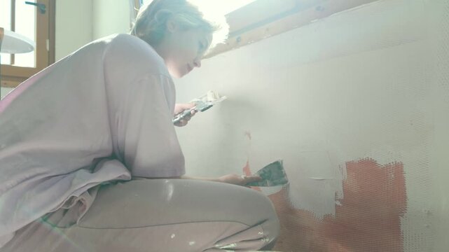 Teenage girl spreading putty across wall surface. Filling cracks with spackle, preparing walls for painting. Helping parents with home renovation. Slow motion sunny bright indoor footage.