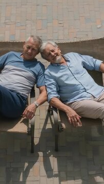 Vertical video: Lying seniors looking up at camera on patio chairs, holding glasses, in blue shirts