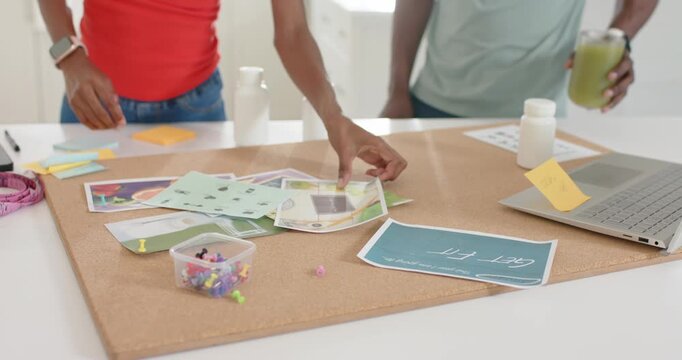 Coworkers woman reaching, arranging pages on corkboard as man holding green drink planning layout