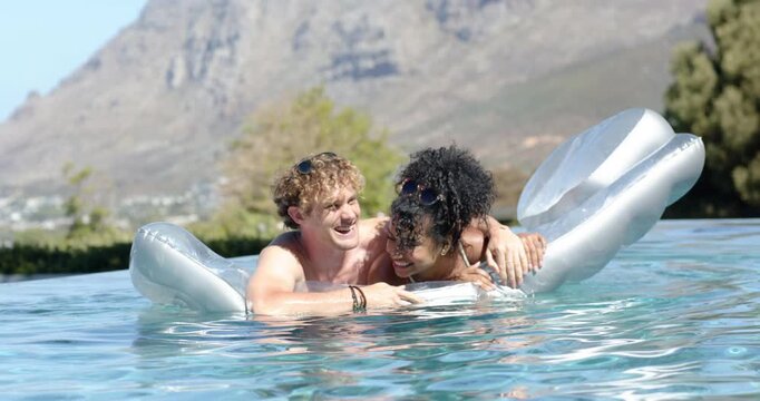 Diverse couple laughing and nuzzling on silver float in pool, man leaning in steadying balance