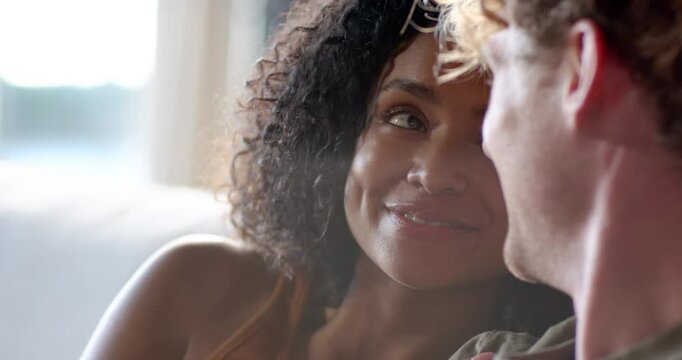 Diverse couple on couch, woman softly speaking in tan top, hand on man's chest, sharing affection