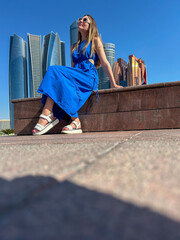 Fototapeta premium Fashionable woman in blue dress sitting on terrace with modern skyscraper skyline, sunny urban travel lifestyle