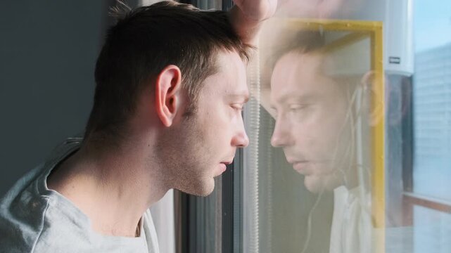 Sad man looking through window with reflection feeling loneliness and depression at home. Thoughtful male in quiet moment of sadness and self reflection, suggesting midlife crisis, emotional stress