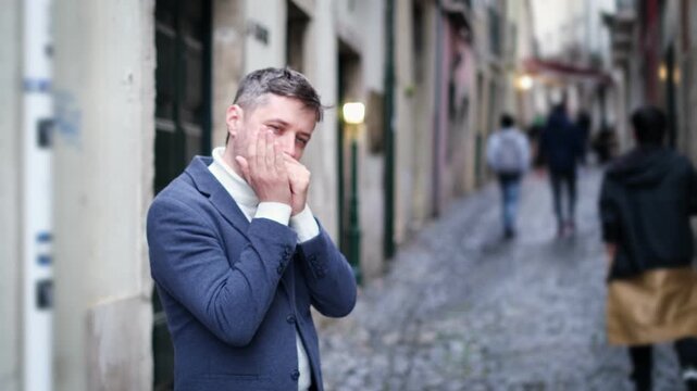 A man plays the harmonica while standing on a narrow street in Lisbon, Portugal. People walk by as he performs, showcasing his musical talent in the city.