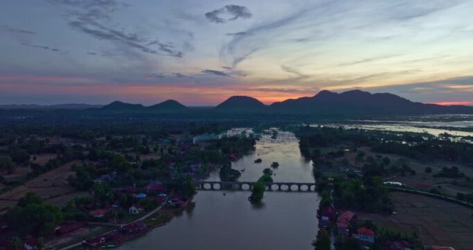 A scenic aerial view of the red bridge crossing the Mekong River near Li Phi Waterfall also known as Tad Somphamit at sunset. The tranquil river landscape with rural farmland and distant mountains
