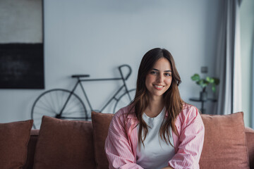 A cheerful young woman sits on a sofa at home, laughing and looking at the camera, with wide copy...