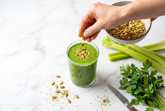 Hand sprinkling pumpkin seeds into green detox smoothie on marble