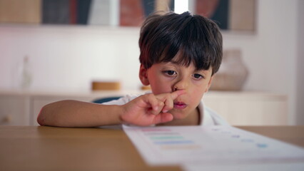 Naklejka premium Young boy struggling to focus on homeschooling worksheet, absentmindedly pointing at paper, feeling bored and distracted while trying to stay engaged in learning at home