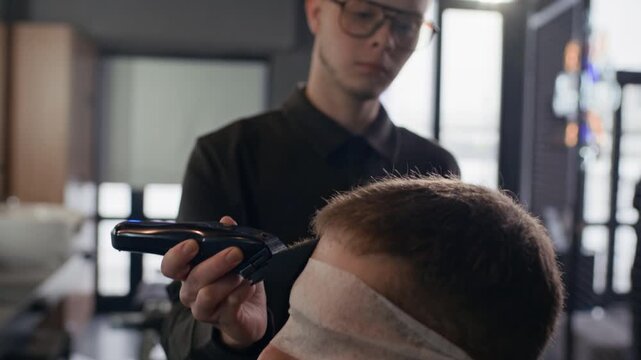 Tilt shot of young Caucasian barber using electric razor to trim client head. Client sitting with closed eyes in barbershop chair