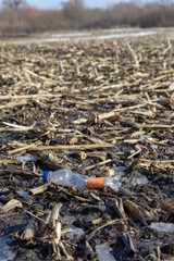 Obraz premium A crushed plastic water bottle with an orange label lies in a muddy field among dry corn stalks and melting ice patches.