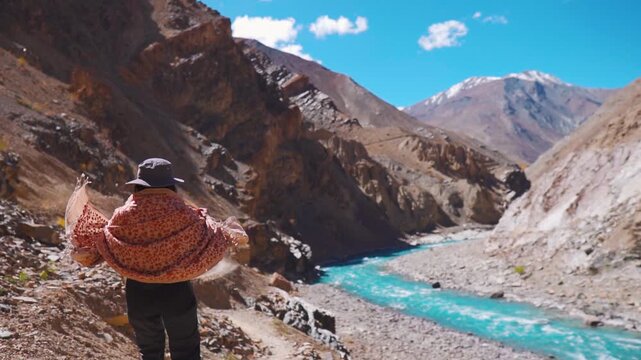 Young Indian woman wearing shawl and cap standing near Blue Tsarap river at Phugtal monastery trek in Zanskar, Ladakh, India. Tourist woman raising hands against Chenab river flowing in valley. Travel
