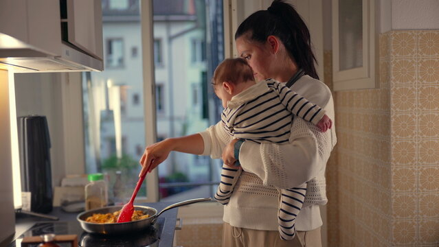 Mother holding baby while cooking in kitchen, stirring food in frying pan, multitasking with child in arms, everyday parenting responsibilities in home setting