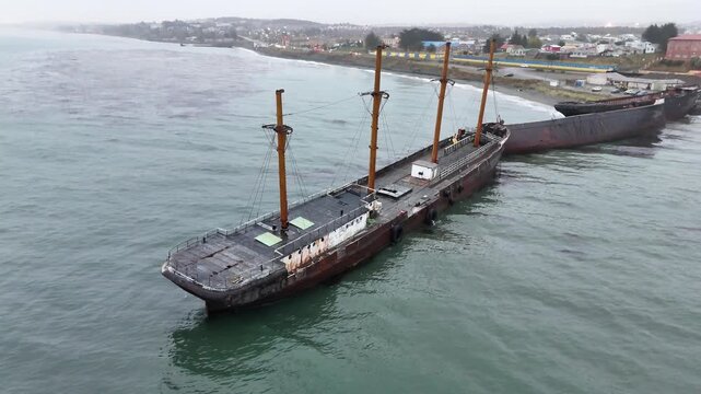 Aerial rises vertically beside the rusted iron hull of the Mu&ntilde;oz Gamero (County of Peebles) shipwreck in Punta Arenas, Chile, revealing its historic masts and the Strait of Magellan