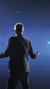 Professional speaker delivering a conference speech on a stage illuminated by spotlights and filled with swirling smoke. Wearing a headset microphone and gesturing with his hands, he engages the