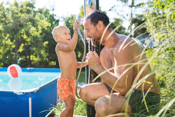 Son and father under shower by the pool on sunny summer day.