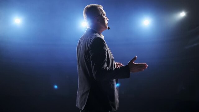 Senior male motivational speaker passionately gestures while delivering an inspiring speech on stage, wearing a headset microphone. Vibrant stage lights illuminate the scene, creating a dynamic and