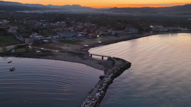 Aerial Drone View of Roda Beach in Corfu Greece, Featuring a Stone Breakwater Jetty, Small Bridge, and Calm Ionian Sea During a Colorful Sunrise Over the Mediterranean Coastline