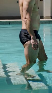 Vertical video: Toe touching, man in black swim briefs stepping down steps at pool to enter water