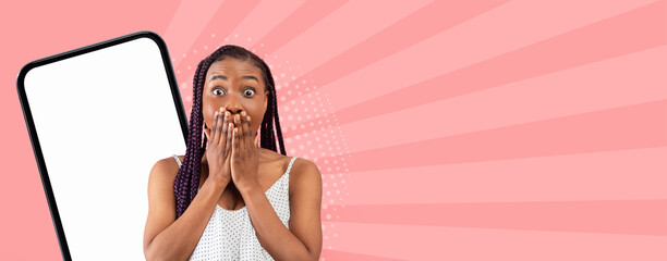A woman stands with her mouth covered in surprise. She is in front of a blank mobile screen. The background features bright colors. The scene shows an expression of shock or excitement. © Prostock-studio
