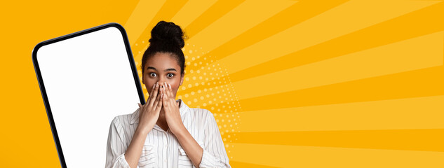 A young woman stands with her hands covering her mouth, showing a look of shock. She is in front of a large phone with a blank screen. The bright yellow background adds energy to the scene. © Prostock-studio
