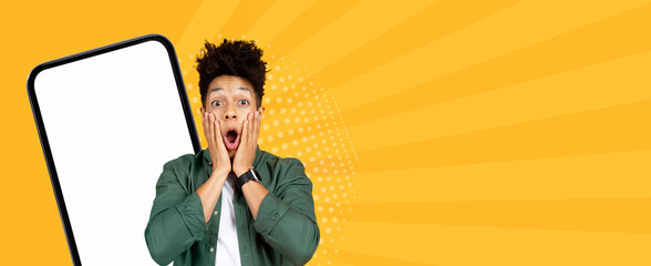 A man stands with wide eyes and hands on his face, expressing surprise. He is in front of a bright yellow background and a blank smartphone screen, making it an engaging visual. © Prostock-studio